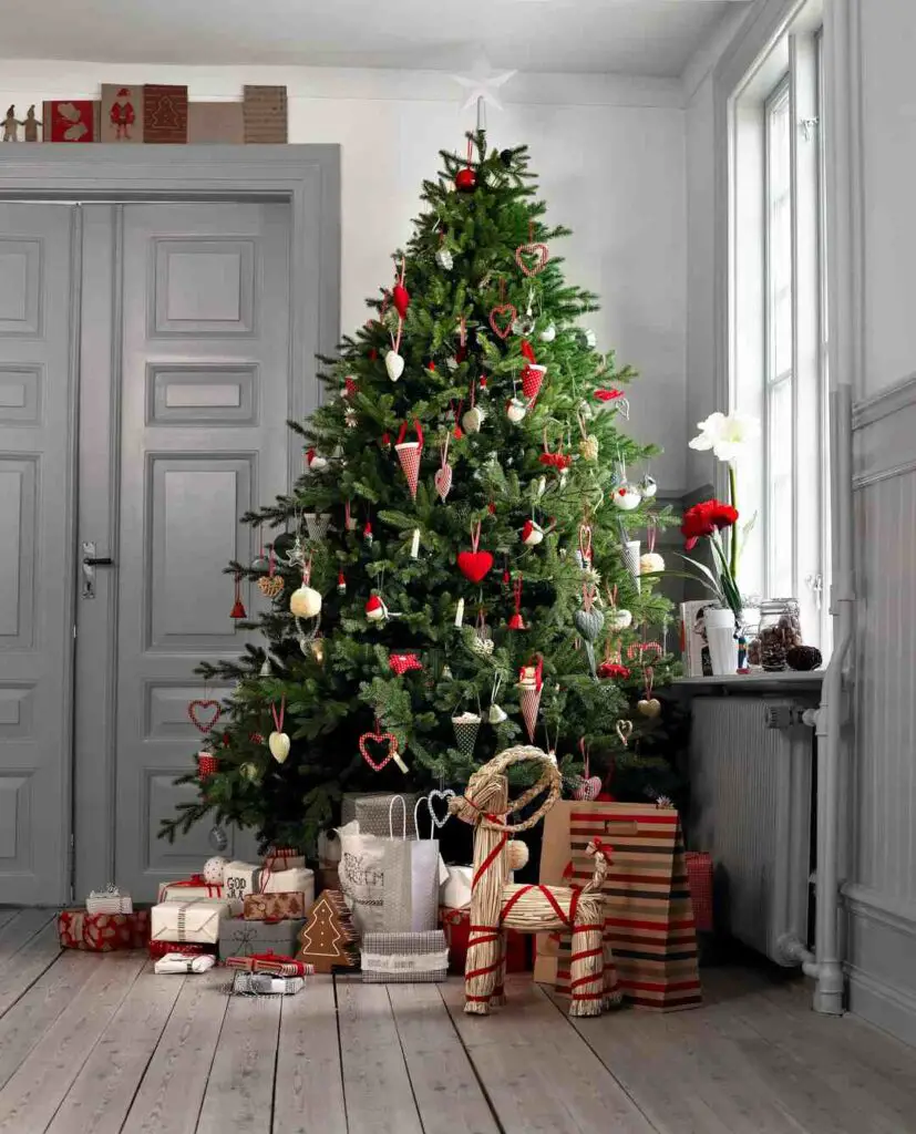  "A beautifully decorated Christmas tree stands tall in a serene room with light gray walls and wooden flooring. The tree is adorned with an array of handmade ornaments, including red hearts, white angels, and various festive baubles. Wrapped presents in coordinating red, white, and natural tones are thoughtfully placed around the tree's base, adding to the warm and inviting holiday atmosphere."