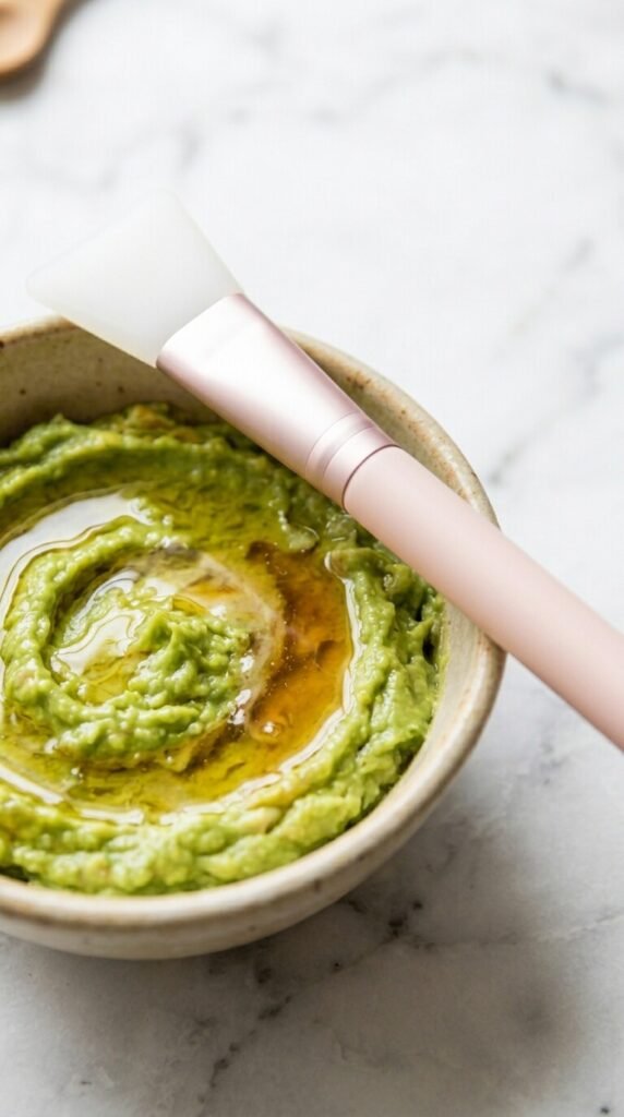 A close-up of an organic DIY avocado face mask with a honey swirl inside a ceramic bowl, featuring a pink silicone application brush resting on top for an at-home spa night.