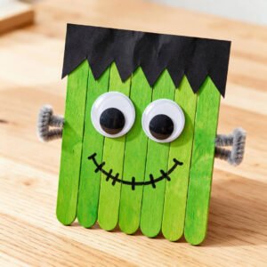 Green painted wooden popsicle stick Frankenstein monster craft with black jagged hair, two googly eyes, and pipe cleaner neck bolts.